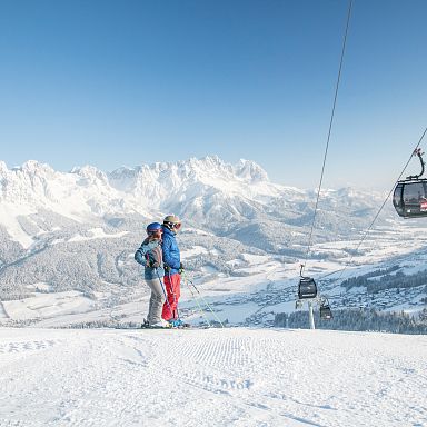 Zwei Skifahrer stehen auf einem schneebedeckten Berg mit Blick auf die Alpen. Im Hintergrund eine Seilbahn und tief verschneite Landschaft unter blauem Himmel.