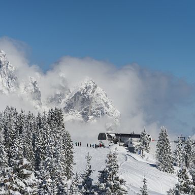 Snowy alpine landscape with pine trees and a ski lodge, surrounded by cloud-covered mountains and a clear blue sky, capturing a wintery scenic view.