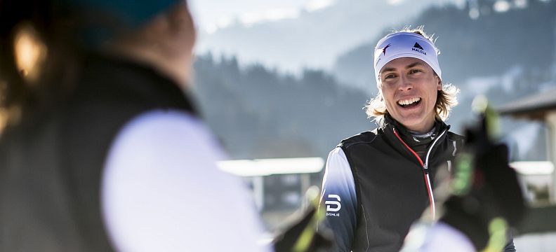 Zwei Personen in Winterkleidung lächeln sich beim Skifahren an. Eine sonnige, schneebedeckte Landschaft mit Bergen im Hintergrund. Winterspaß und Freude.