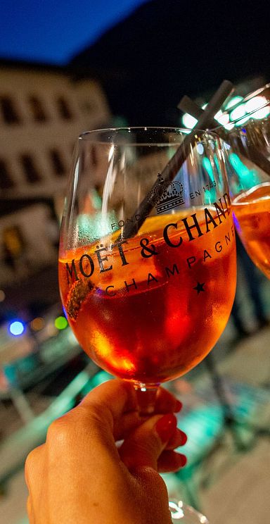 Zwei Personen stoßen mit gefüllten Weingläsern an, im Hintergrund spielt ein Musiker Gitarre vor einem beleuchteten Gebäude am Abend.