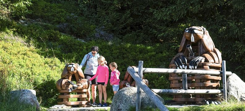 Familie betrachtet Holzskulpturen von Murmeltieren in einer grünen, bewaldeten Umgebung. Der sonnige Tag hebt die natürliche Landschaft hervor.