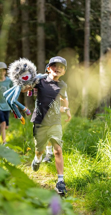 Children running joyfully on a forest trail in the Wilder Kaiser region, surrounded by lush greenery and sunlight peeking through the trees.