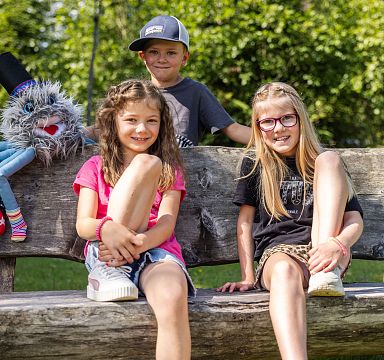 Three children sitting on a wooden bench outdoors, smiling, with a colorful puppet beside them. Trees and greenery can be seen in the background.