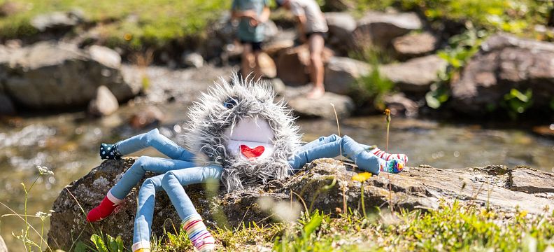 A colorful plush spider toy on a rock by a stream, with children playing nearby in the Wilder Kaiser region. Sunny day in a lush, green outdoor setting.