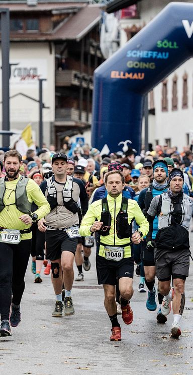 Gruppe von Läufern mit Startnummern, darunter einige mit Stirnlampen, starten bei einem Event in der Bergregion Wilder Kaiser, unter einem großen blauen Tor.