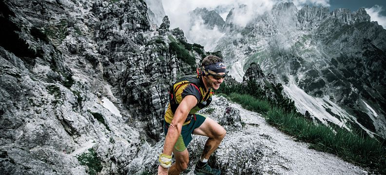 A hiker in sports gear climbs steep, rocky terrain in the Wilder Kaiser mountains, with dramatic peaks and clouds in the background, showcasing outdoor adventure.