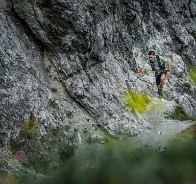 A hiker climbs on rocky terrain at Wilder Kaiser, surrounded by rugged landscape and green foliage.