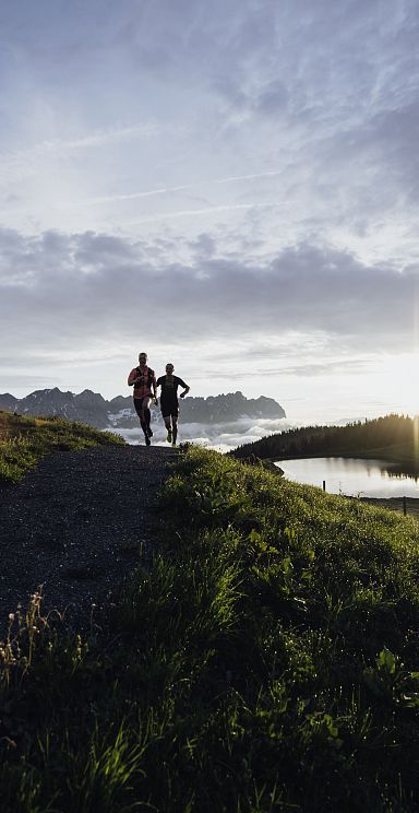 Two people run on a path in the Wilder Kaiser region at sunrise. The sun rises over the mountains, casting light on the grassy landscape and a small pond.