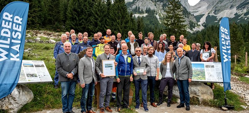 A group of people standing in front of the Wilder Kaiser mountains, with banners and awards, capturing a moment in the scenic Tyrolean setting.