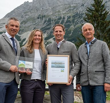 Group receiving an award at Wilder Kaiser with mountains in the background. They are dressed in traditional attire and smiling, surrounded by scenic nature.