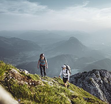 Zwei Wanderer erklimmen einen grasbewachsenen Hang mit felsigen Ausläufern im Hintergrund, während die Sonne über den Alpen aufgeht und Nebel die Täler füllt.