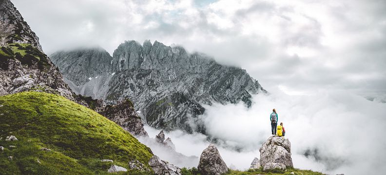 Two hikers on a rocky ledge in the Wilder Kaiser, amidst green slopes and mist-covered peaks, epitomize the grandeur of this alpine landscape.