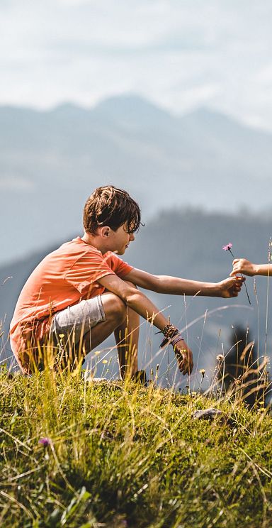 Two children, a boy and a girl, joyfully play on a mountain meadow in Wilder Kaiser. They seem to be exchanging something in the vibrant green landscape.