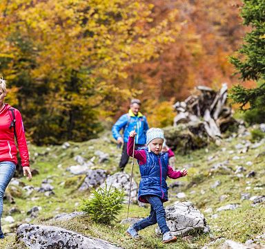 A family hikes through Wilder Kaiser in autumn, with colorful leaves and rocky terrain. A child leads the way, showcasing the beauty of the region.