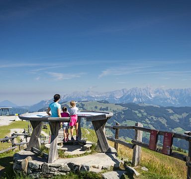 Zwei Menschen betrachten von einem Aussichtspunkt aus die Alpenlandschaft mit grünen Wiesen und Bergen unter klarem, blauem Himmel in Tirol.