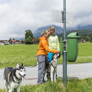 Two people with dogs stand by a green field with the Wilder Kaiser mountains in the background and a trash bin nearby.