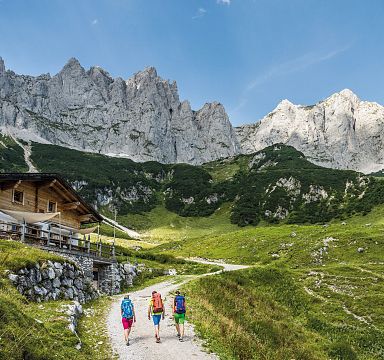 Three hikers walk on a mountain path near a hut, with the towering rocky peaks of the Wilder Kaiser in the background under a clear blue sky.