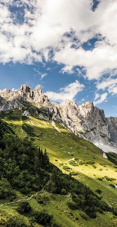 Scenic view of the Wilder Kaiser mountains with green hills, rocky peaks, and blue sky with clouds, ideal for hiking and nature lovers.