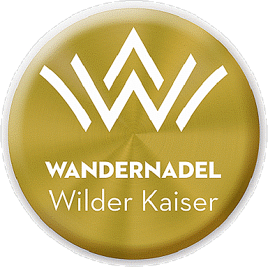 Graphic of the Wilder Kaiser hiking badge with a stylized mountain logo and the words 'Wandernadel Wilder Kaiser' on a gold circular background.