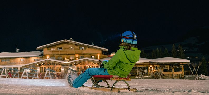 A child on a sled enjoys nighttime sledding near the Wilder Kaiser. The area is lit by lights from a nearby lodge, adding a warm and inviting feel.