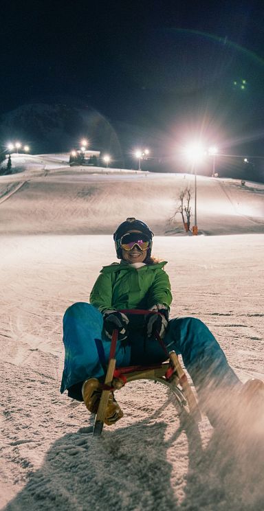 A person sledding down a well-lit snowy hill at night in Wilder Kaiser. The scene captures a thrilling ride with illuminated ski slopes in the background.