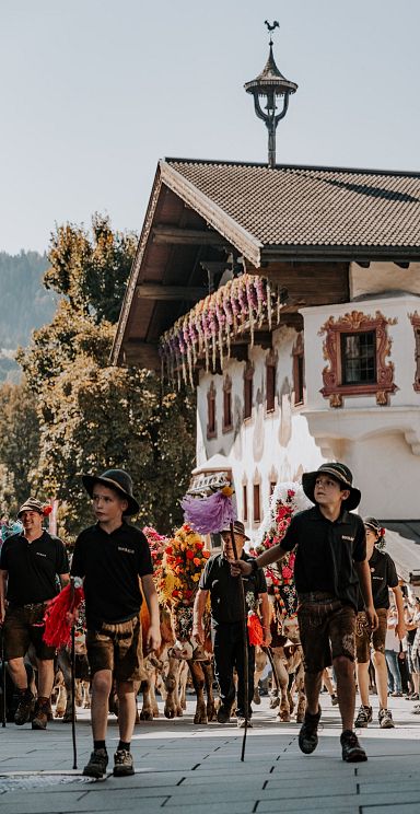 Kinder in traditionellen Hüten laufen vor einem verzierten, alpenländischen Haus mit buntem Blumenschmuck und dekorativen Wandmalereien.