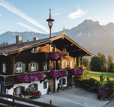 Traditionelles Tiroler Holzhaus mit blühenden Geranien, im Hintergrund das beeindruckende Bergmassiv des Wilden Kaisers bei Sonnenaufgang.