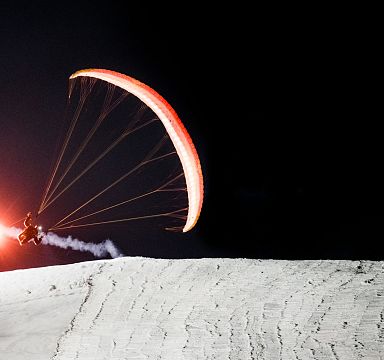 Ein Paraglider fliegt nachts über verschneite Hänge, beleuchtet von einer roten Leuchtfackel, die einen faszinierenden Lichtschweif hinterlässt.