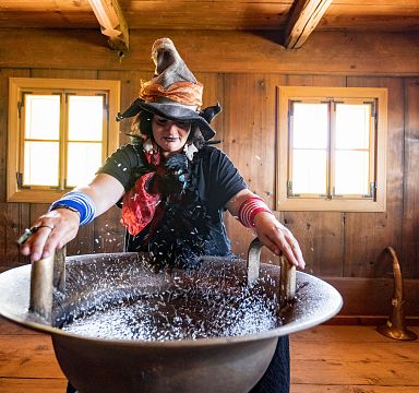 Frau in traditioneller Kleidung mit Hut steht in rustikaler Holzhütte und streut Körner in eine große Schale. Die Szene wirkt traditionell und authentisch.