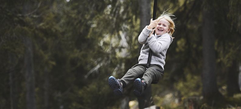 A young child swings joyfully in a forest setting near the Wilder Kaiser mountains, surrounded by trees and a sense of adventure and fun.