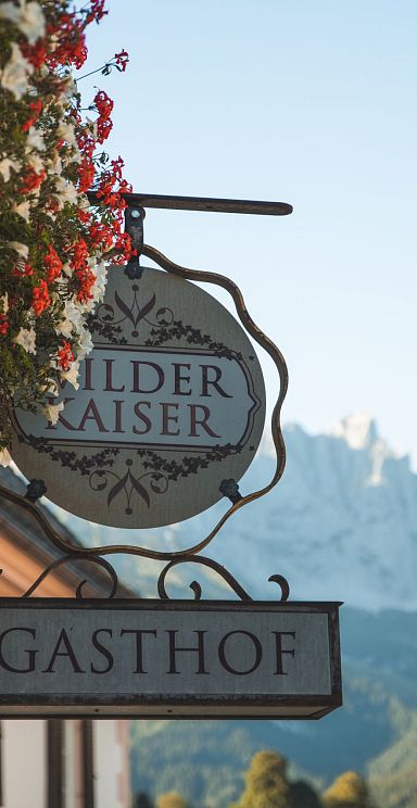 Gasthof-Schild mit "Wilder Kaiser"-Schriftzug, umliegende Alpenlandschaft und Berge im Hintergrund bei klarem Wetter, rustikaler Charme.