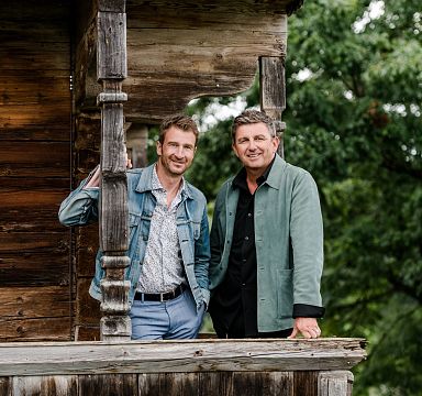 Zwei Männer stehen auf einer Holzveranda vor einem rustikalen Gebäude. Sie lächeln in die Kamera. Im Hintergrund sind Bäume zu sehen.