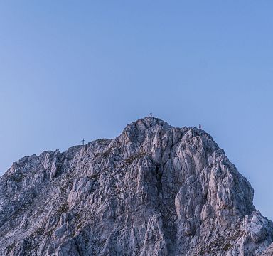 Ein felsiger Berggipfel unter klarem blauem Himmel mit mehreren Gipfelkreuzen. Die Felswände haben eine markante, zerklüftete Struktur.