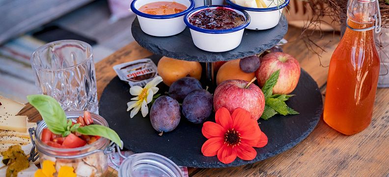Ein Frühstückstisch mit Obst, Marmelade, Brotaufstrichen, einem Getränk in einer Glasflasche und Blumen dekoriert. Alles ist appetitlich angerichtet.