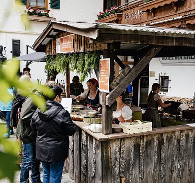 Menschen stehen an einem rustikalen Marktstand aus Holz, dekoriert mit hängenden Kräutern, während traditionell gekleidete Verkäuferinnen Speisen servieren.