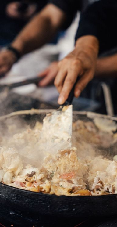 Nahaufnahme von Händen, die ein Gericht in einer Pfanne mit einem Spatel zubereiten. Dampf steigt auf, während das Essen gerührt wird.