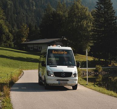 White shuttle bus on a narrow mountain road surrounded by lush greenery in the Wilder Kaiser area.