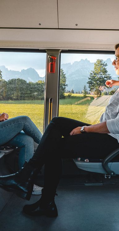 Two individuals talking inside a bus with the majestic Wilder Kaiser mountains visible outside through the window, showcasing a scenic backdrop.