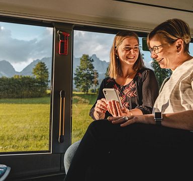 Two women sit inside a train, smiling and looking at a smartphone. Outside the window, the Wilder Kaiser mountains rise against a clear, sunny sky.