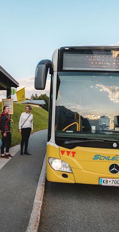 Tourists wait at a scenic bus stop in the Wilder Kaiser region, surrounded by greenery and distant mountains. A yellow bus is parked at the roadside stop.