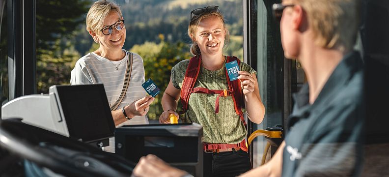 Zwei Personen kaufen Fahrkarten bei einem Busfahrer, während sie lächeln und sich unterhalten. Im Hintergrund sind Bäume und Berge zu sehen.