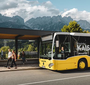 A yellow bus at a stop with people walking nearby, set against the backdrop of the Wilder Kaiser mountains and a partly cloudy sky.