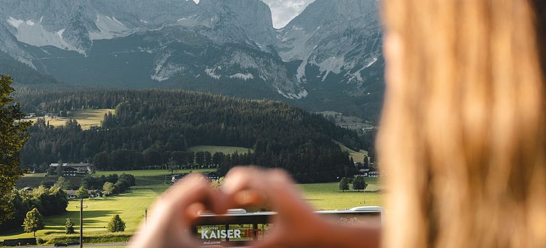 View of the Wilder Kaiser mountains with green fields and a person making a heart shape with hands in the foreground.