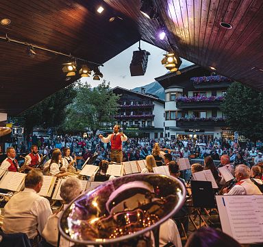 Eine Blaskapelle spielt unter einem Holzdach bei einem Freiluftkonzert. Zahlreiche Zuhörer sind vor traditionellen Tiroler Häusern versammelt.