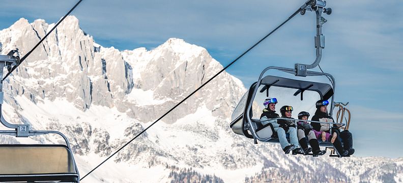 Skilift mit mehreren Personen vor schneebedecktem Bergmassiv des Wilden Kaisers unter blauem Himmel. Winterliches Alpenpanorama in Tirol.