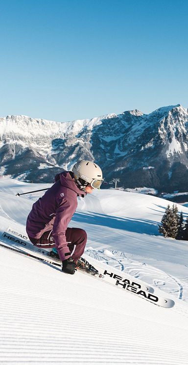 Skier in a purple jacket descends snowy slopes at Wilder Kaiser with mountain range in the background under a clear blue sky.