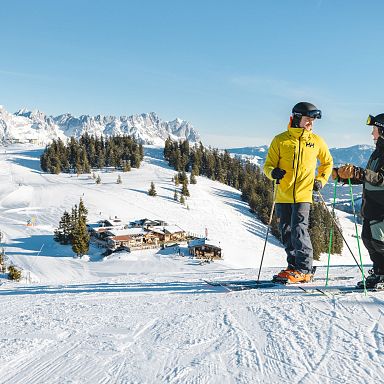 Zwei Skifahrer stehen auf einer verschneiten Piste mit Blick auf schneebedeckte Berge. Im Hintergrund sind Hütten und Bäume zu sehen, der Himmel ist klar.
