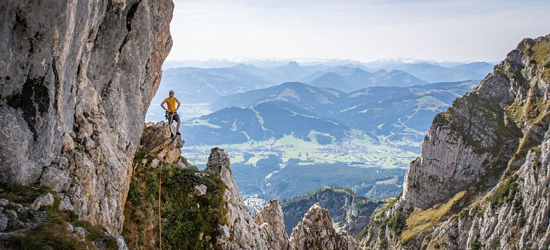 A hiker on a rocky outcrop in the Wilder Kaiser Mountains with expansive views of valleys and distant hills under a clear sky.