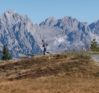 A runner on a grassy trail with the stunning peaks of the Wilder Kaiser mountain range in the background. Sparse pine trees and clear skies complete the scenic view.