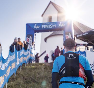 Ein Läufer in Sportkleidung nähert sich dem Ziel eines Berglaufs, flankiert von Zuschauern. Im Hintergrund ist eine Kapelle bei Sonnenschein zu sehen.
